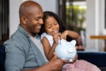 Father and daughter sitting on the couch with a piggy bank