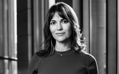 Black and white headshot of a person with shoulder-length hair, standing in a modern indoor setting with large windows in the background.