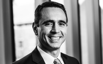 BW FacultyPhotos Joao Gomes Black and white headshot of a person in a suit and tie, smiling, with a blurred background of windows.
