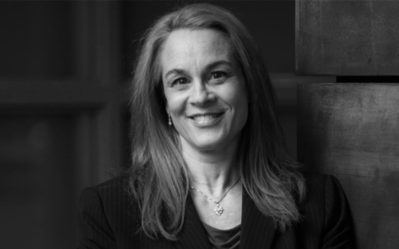 A black and white headshot of a smiling person with long hair, wearing a suit jacket and a necklace.