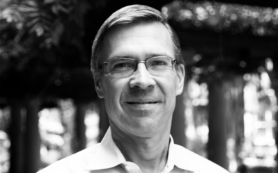 BW FacultyPhotos Witold Henisz Black and white headshot of a person wearing glasses, standing outdoors. The background is blurred and features some foliage.