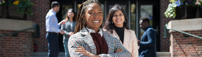 A confident individual with braided hair stands in the foreground, smiling, while a diverse small group of people converses in the background outside a building with steps and flowers.