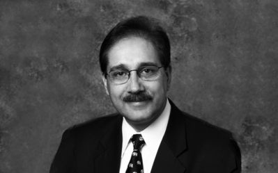 Headshot of a person wearing glasses, a suit, and a patterned tie against a textured background.